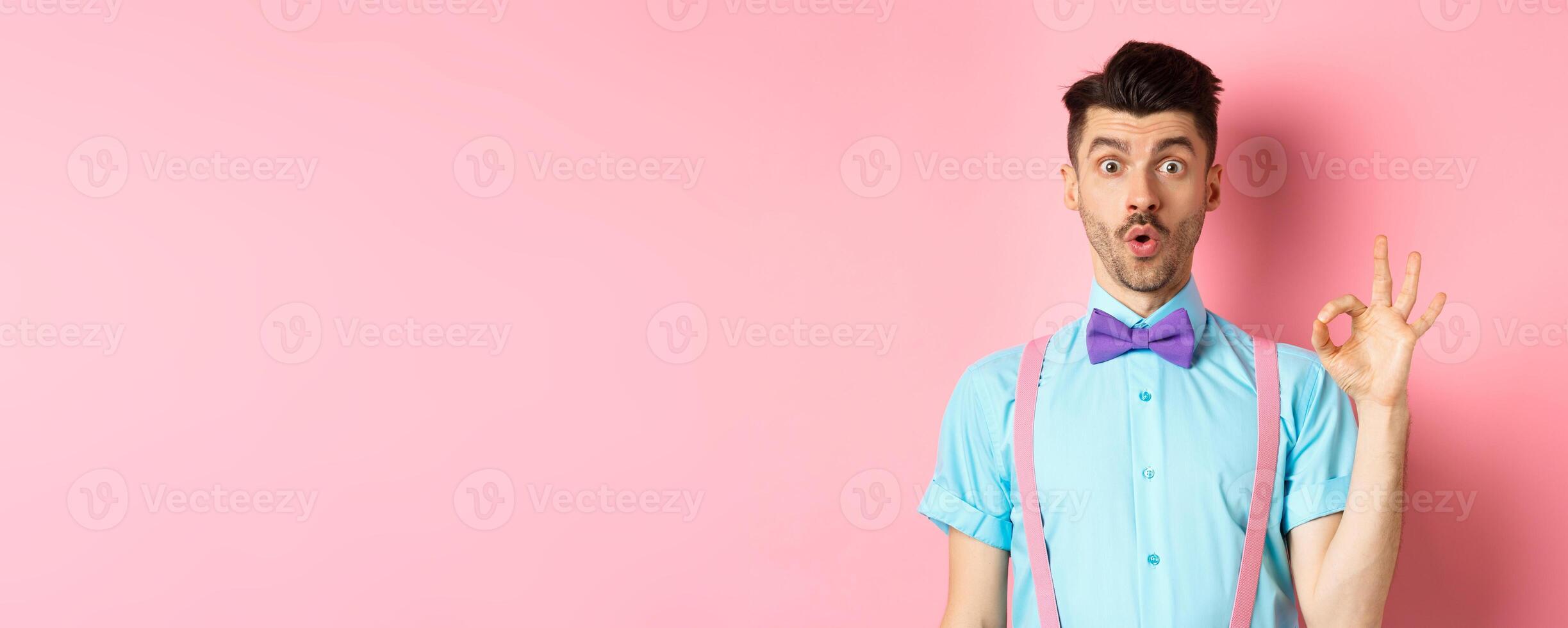 Impressed guy in bow-tie and suspenders saying wow, showing okay gesture with amazement, praise awesome thing, standing over pink background photo