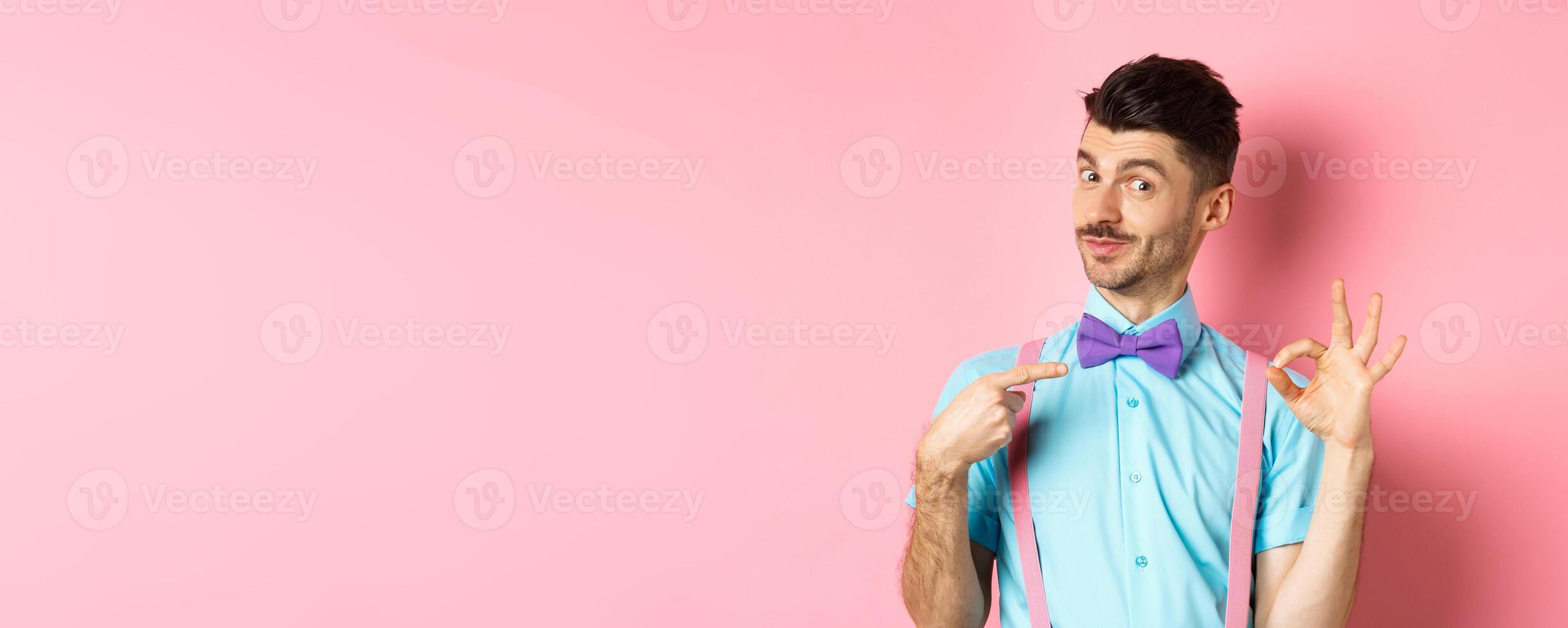 Image of young handsome guy in bow-tie and holiday outfit, pointing at himself and showing okay gesture, self-promoting on pink background photo