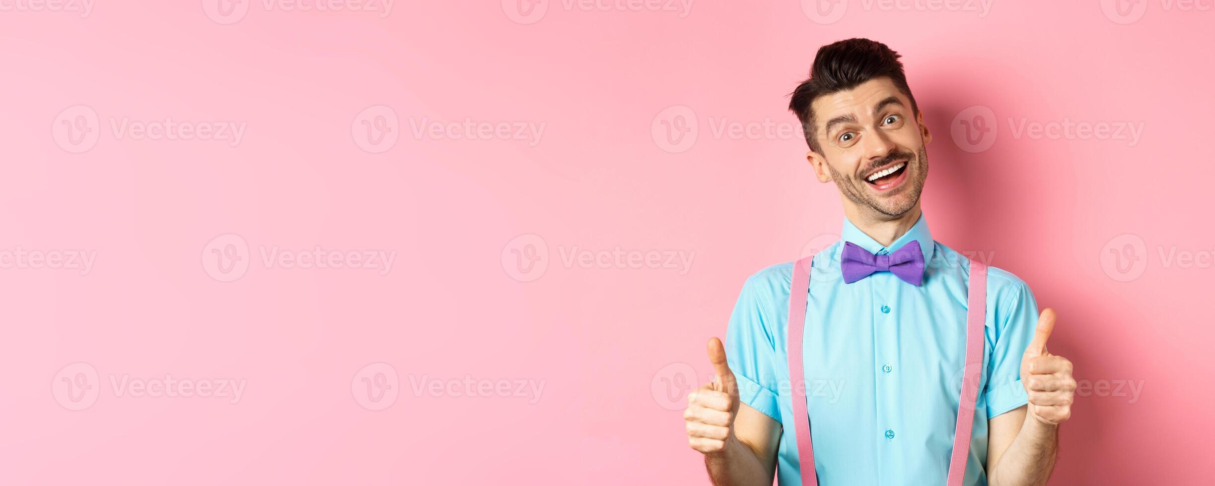 Happy smiling guy in bow-tie and suspenders showing thumbs up, praising good job, approve nice work, standing over pink background satisfied photo