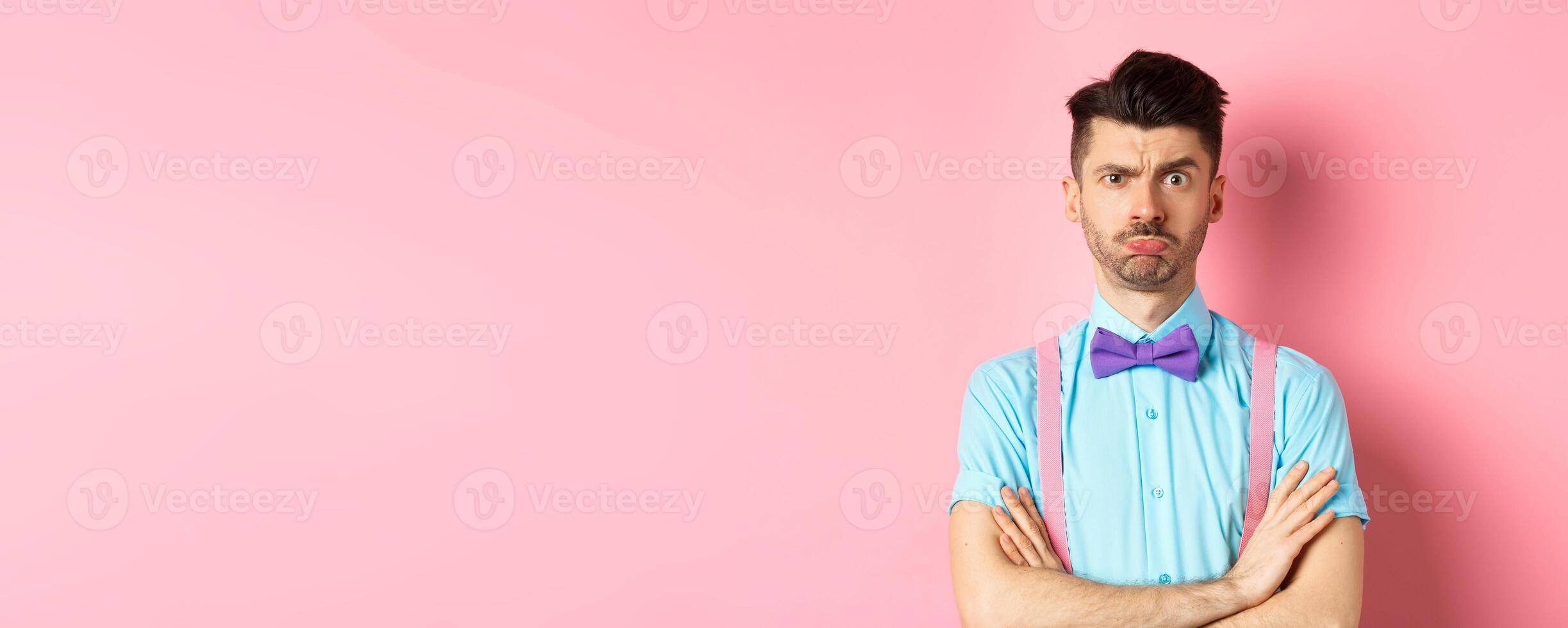 Silly young guy with moustache, sulking and feeling timid, cross arms on chest, being offended at you, standing insulted on pink background photo