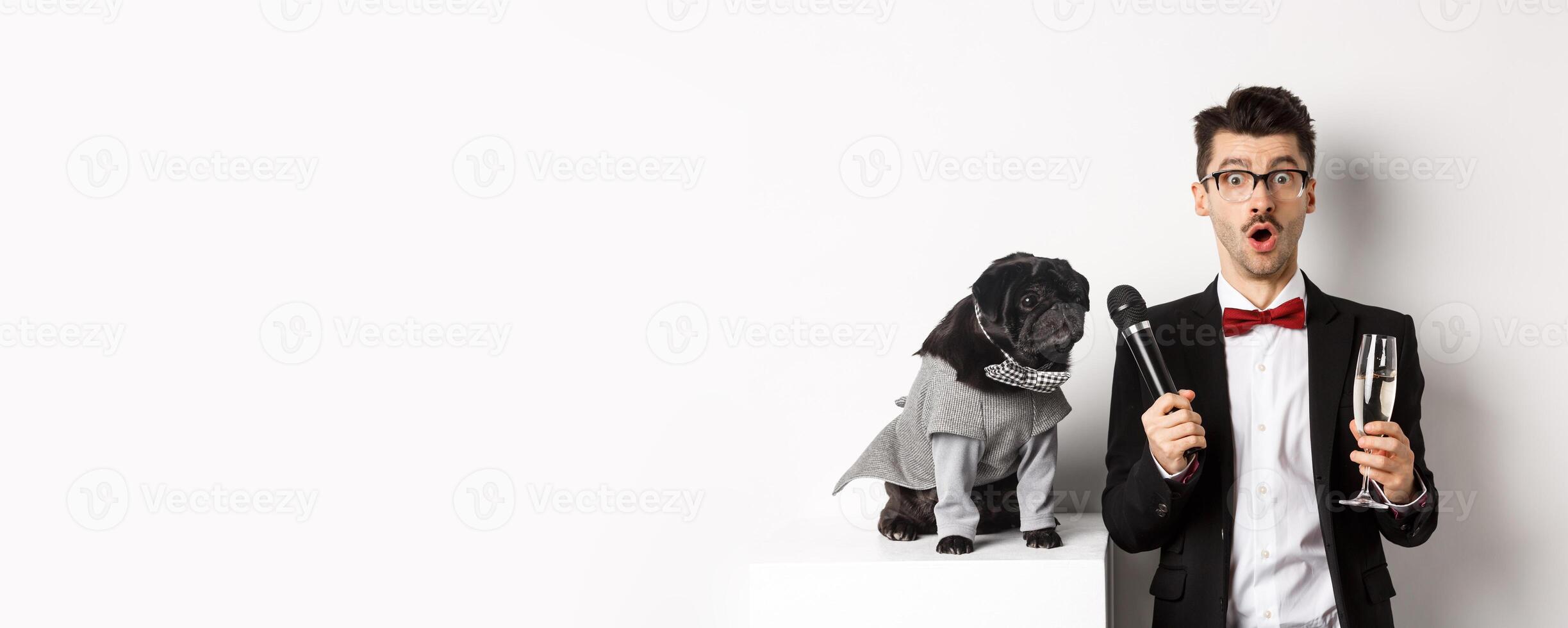 Male entertainer raising glass of champagne, giving microphone to cute black dog, standing over white background photo