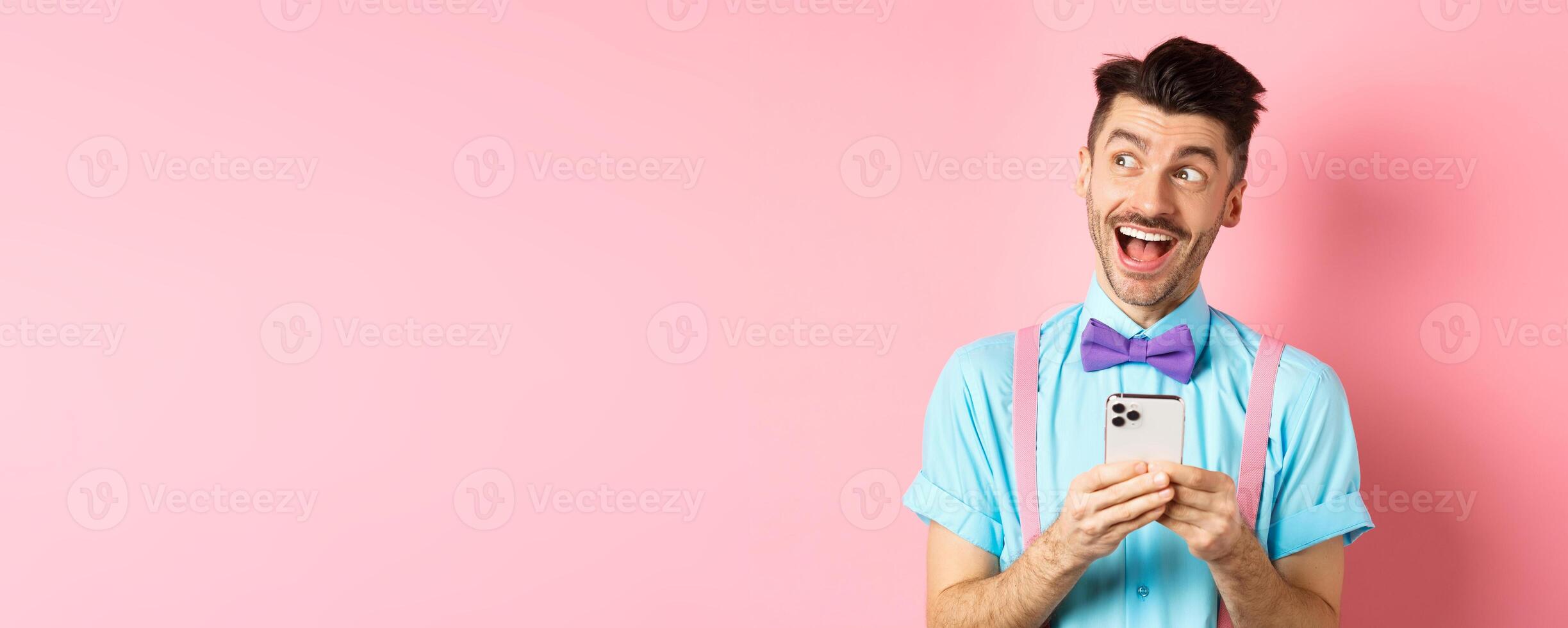 Technology concept. Excited man looking aside with dreamy face, winning online prize on smartphone, standing cheerful over pink background photo