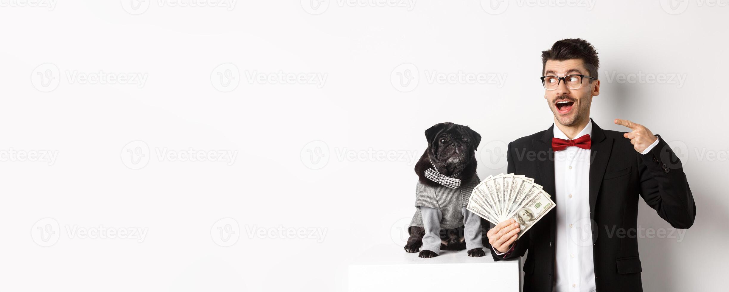 Happy young man in suit earn money with his dog. Guy rejoicing, holding dollars and pointing left, black pug in costume staring at camera, white background photo