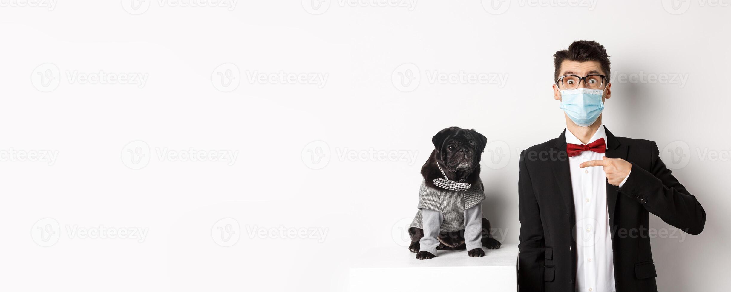 Coronavirus, pets and celebration concept. Amazed young man in face mask and suit pointing at cute black dog sitting near owner in party outfit, white background photo