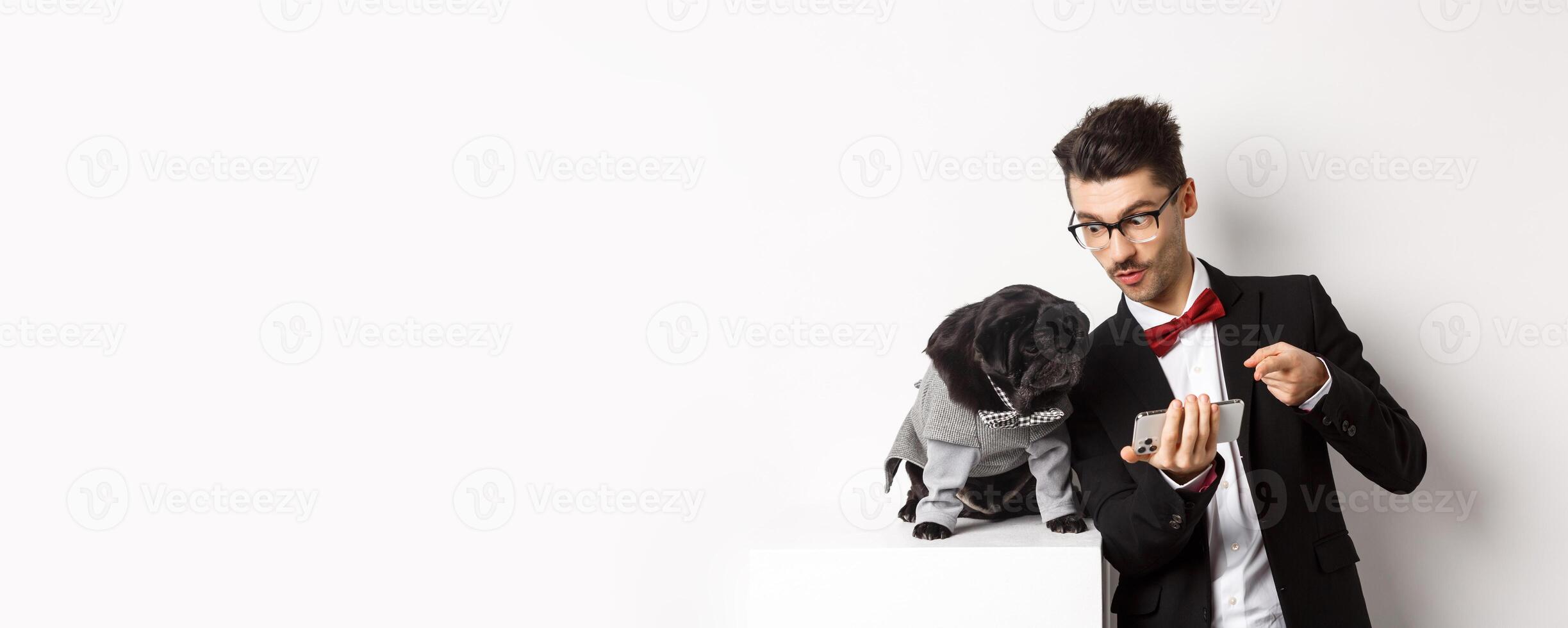Handsome young man showing something on mobile phone to his dog. Owner shopping online with pet, standing in costumes over white background photo