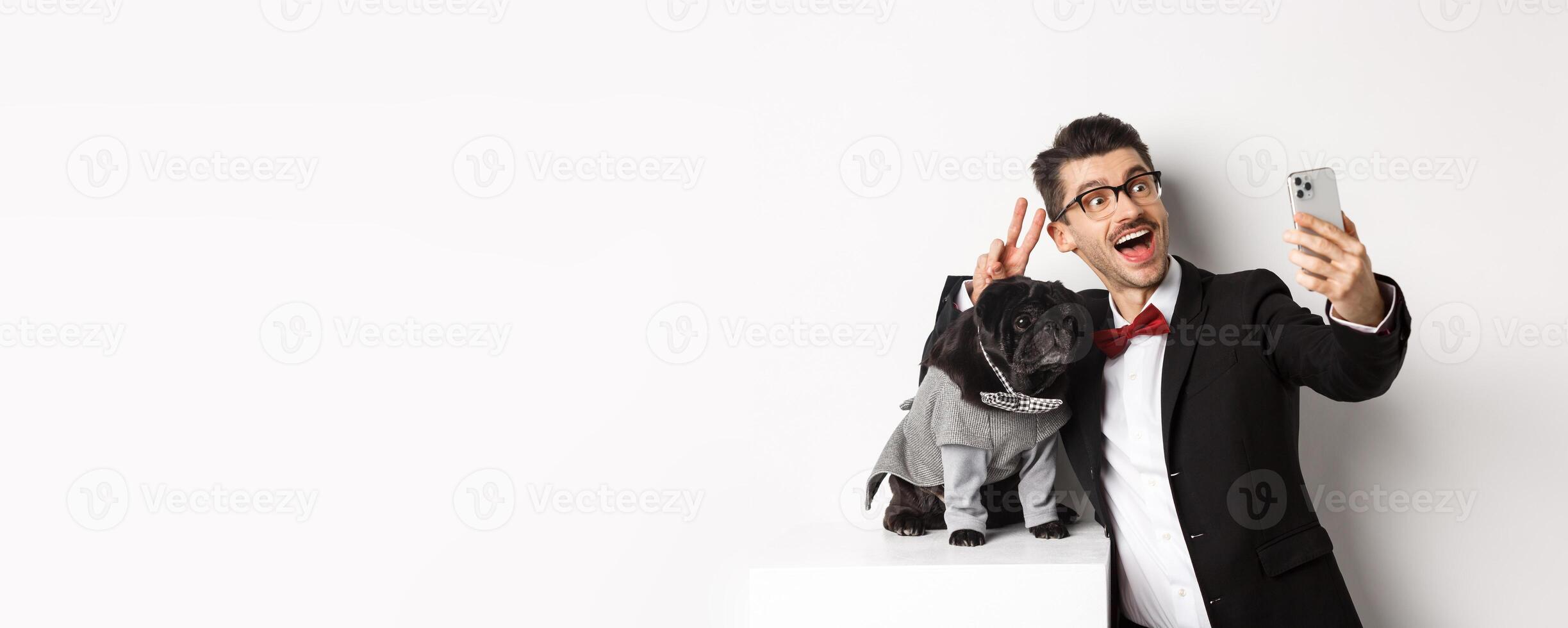 Cheerful dog owner in suit celebrating New Year with dog, taking selfie on smartphone near cute black pug in costume, white background photo