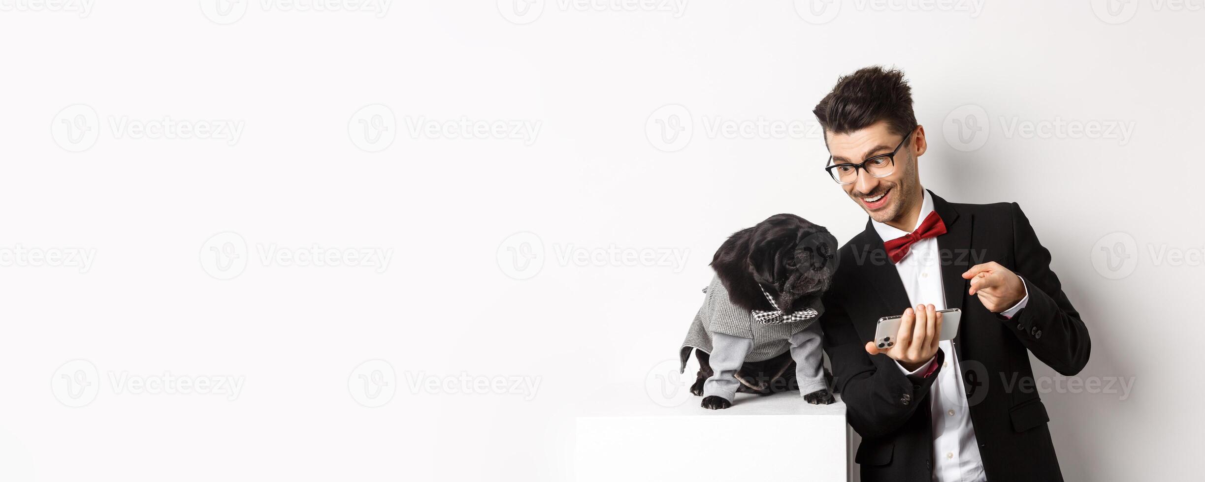 Handsome young man showing something on mobile phone to his dog. Owner shopping online with pet, standing in costumes over white background photo