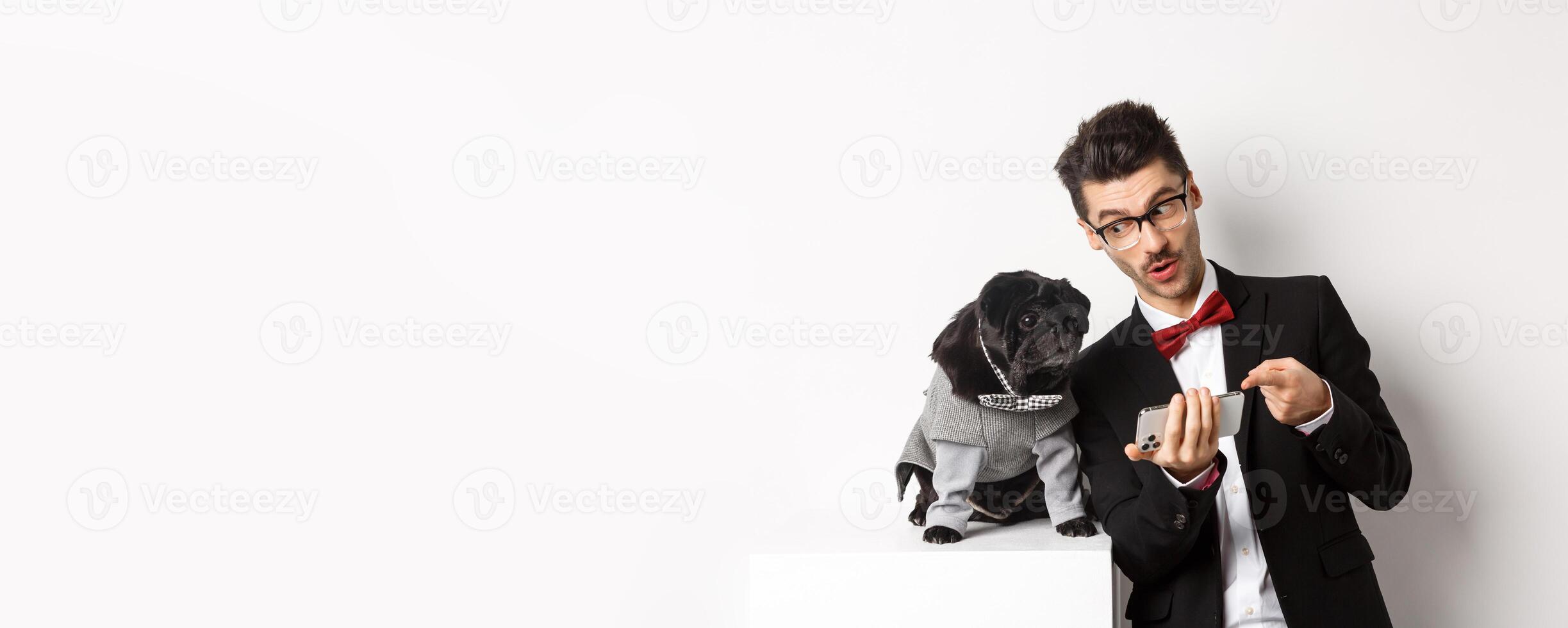 Handsome young man showing something on mobile phone to his dog. Owner shopping online with pet, standing in costumes over white background photo