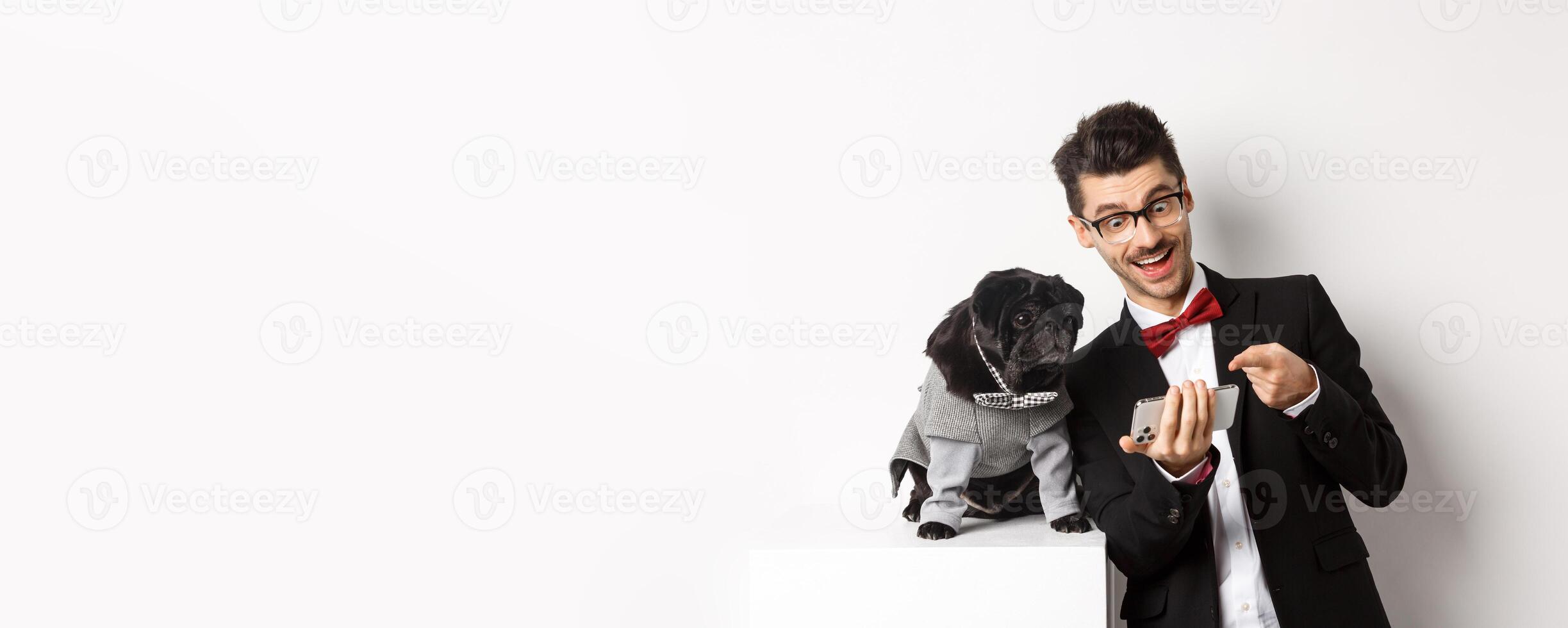 Happy dog owner showing something to pet on mobile phone, man and pug wearing fancy costumes, standing over white background photo