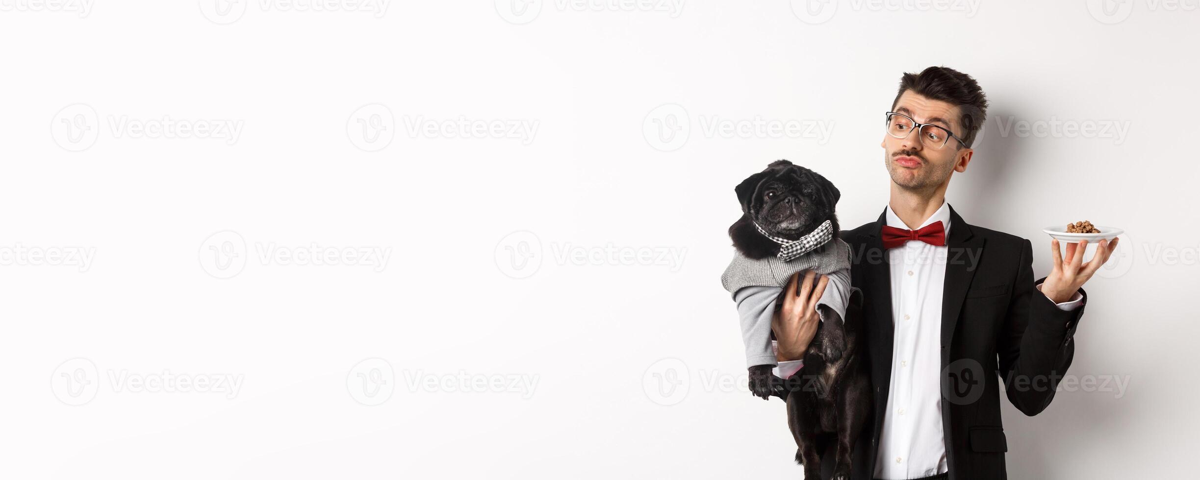Handsome young dog owner in fancy suit holding cute black pug and plate with animal food, standing over white background photo