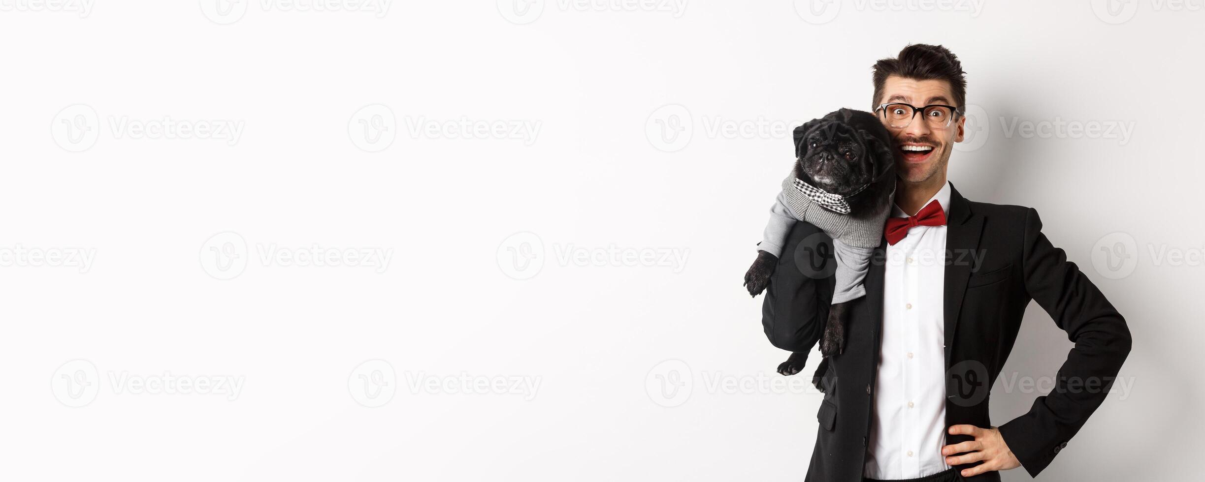Handsome young man in suit and glasses holding cute black pug dog on shoulder, smiling happy at camera, wearing party outfits, white background photo