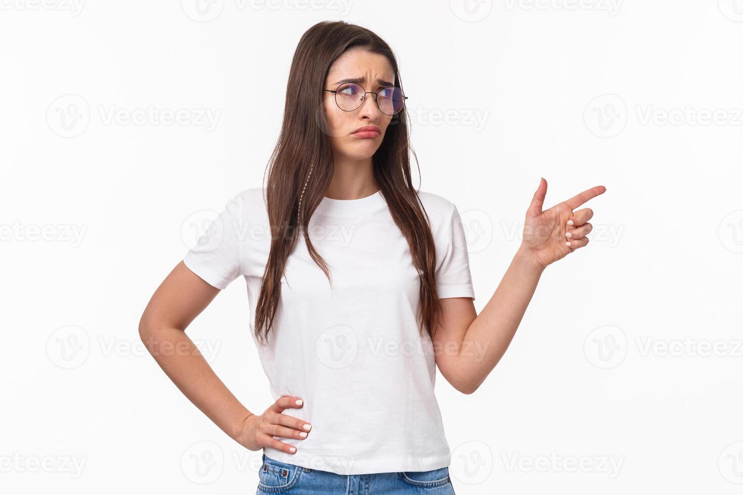 Waist-up portrait of uneasy, sad and gloomy young brunette woman in glasses, looking and pointing finger right with sulking unhappy face, express jealousy or regret, grieving white background photo