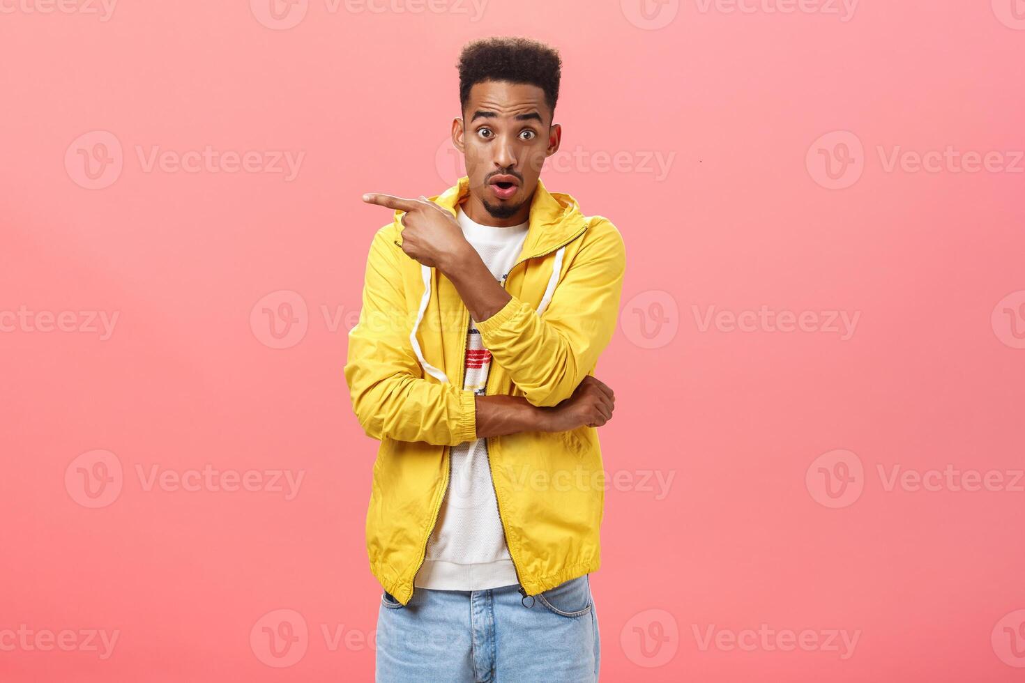 Portrait of impressed good-looking african american male with beard and curly haircut raising eyebrows opening mouth pointing left with questioned shocked expression posing over pink wall photo