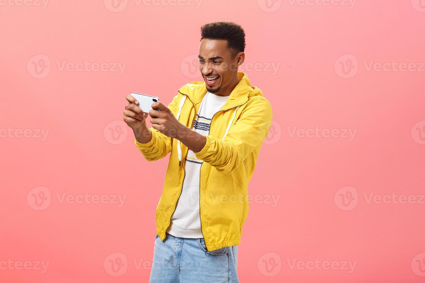 Man being carried away with cool smartphone game holding cellphone with both hands pointing at device screen staring in gadget with intense amazed expression posing over pink background photo