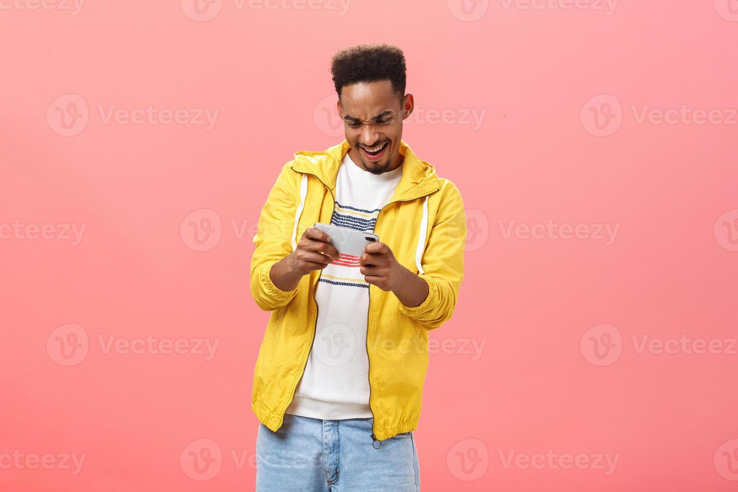 Enthusiastic guy having fun playing awesome smartphone game holding cellphone in both hands staring at device screen with passion and thrill spending free time in internet over pink background photo