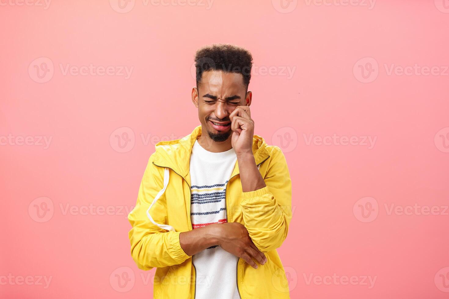Studio shot of silly sad african american timid guy in yellow trendy jacket crying heart out whiping tears from eyes and whining feeling lonely after being rejecting by ex-girlfriend over pink wall photo