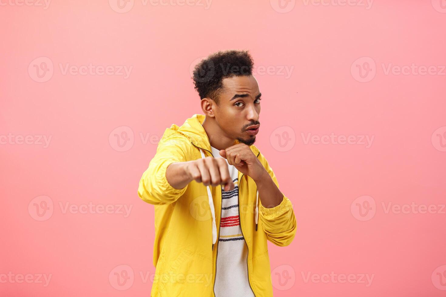 Guy acting cool bragging in front of girlfriend with mascules showing boxer moves standing in boxing pose with raised fist in punch gesture turning right as if fighting over pink background photo