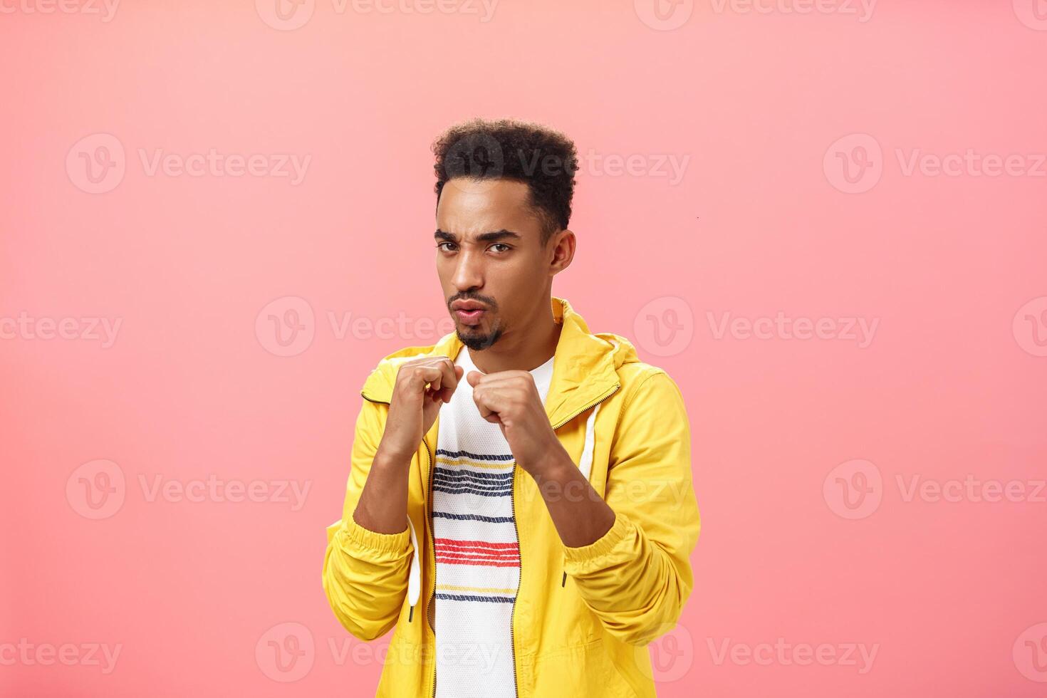 Always ready for battle. Portrait of confident stylish and awesome dark-skinned bearded male with afro haircut raising fists standing in fighting pose being daring and self-assured over pink wall photo