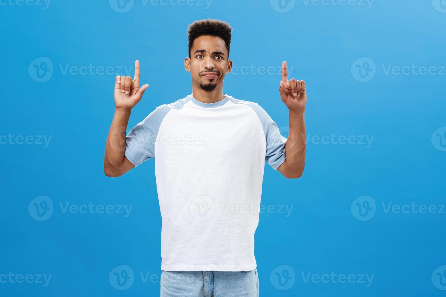 Portrait of displeased intense and doubtful african american male model in casual t-shirt smirking and frowning from disappointment and doubt pointing up seeing something suspicious over blue wall photo