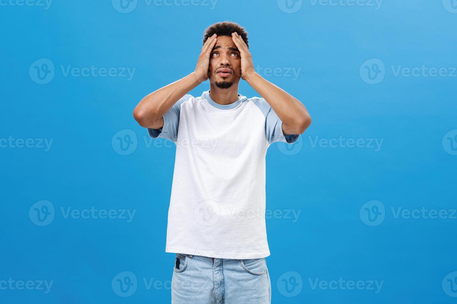 Man feeling reckelss cannot understand why bad things happen him. Distressed panicking and anxious miserable dark-skinned young man holding hands on head looking up in sky being in tough situation photo