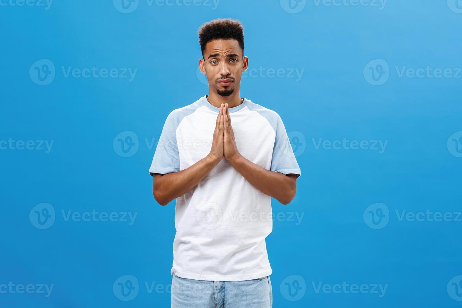 Please help me beg you. Charming cute and handsome african american boyfriend with afro curly haircut and beard holding hands in pray frowning making silly face while asking favor over blue wall photo
