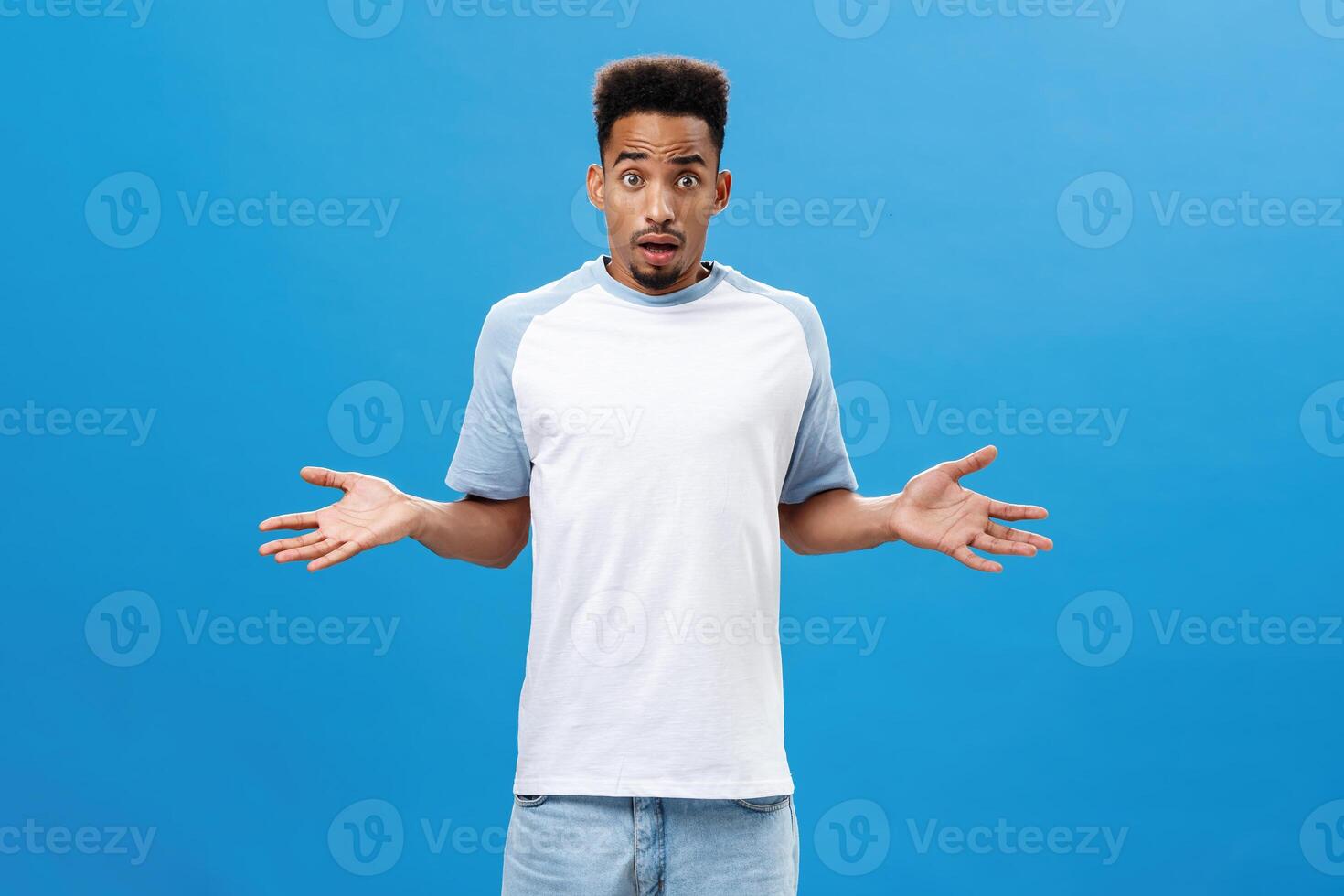 Handsome intense dark-skinned guy feeling concerned and questioned having no clue what happened being worried spreading palms aside with confused expression standing uncertain over blue background photo