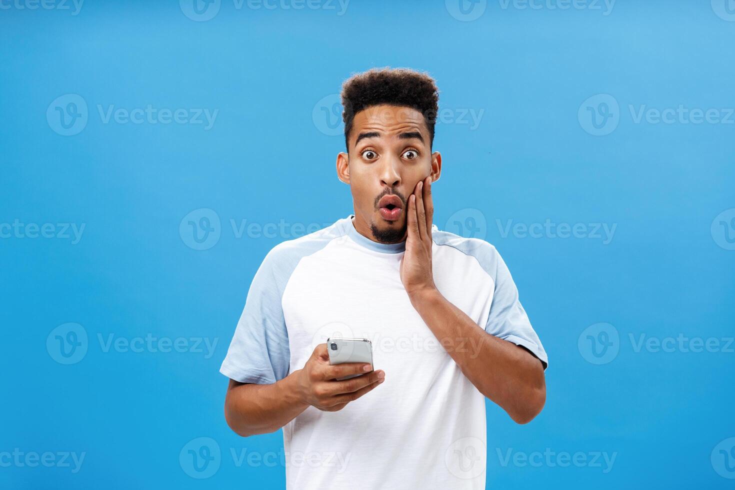 Waist-up shot of charmed intrigued handsome stylish dark-skinned male with afro haircut folding lips in wow or awesome expression holding cheek on face holding smartphone reacting to amazing news photo