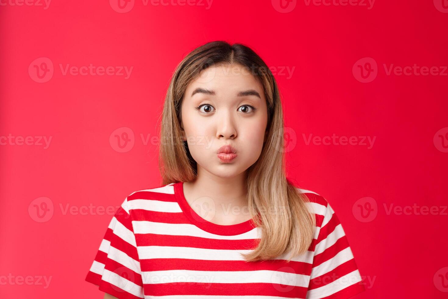 Close-up silly cute asian blond girl pouting making playful face, fold lips hold breath stand red background speechless, stay silent posing childish carefree look impressed photo