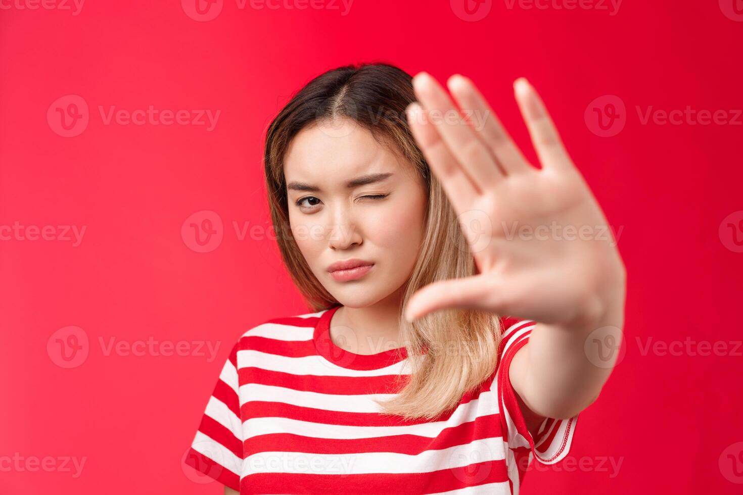 Close-up serious-looking determined asian blond woman pull hand forward show stop taboo sign, concentrating gaze on camera look with one eye, demanding stop, tell enough, say no, red background photo