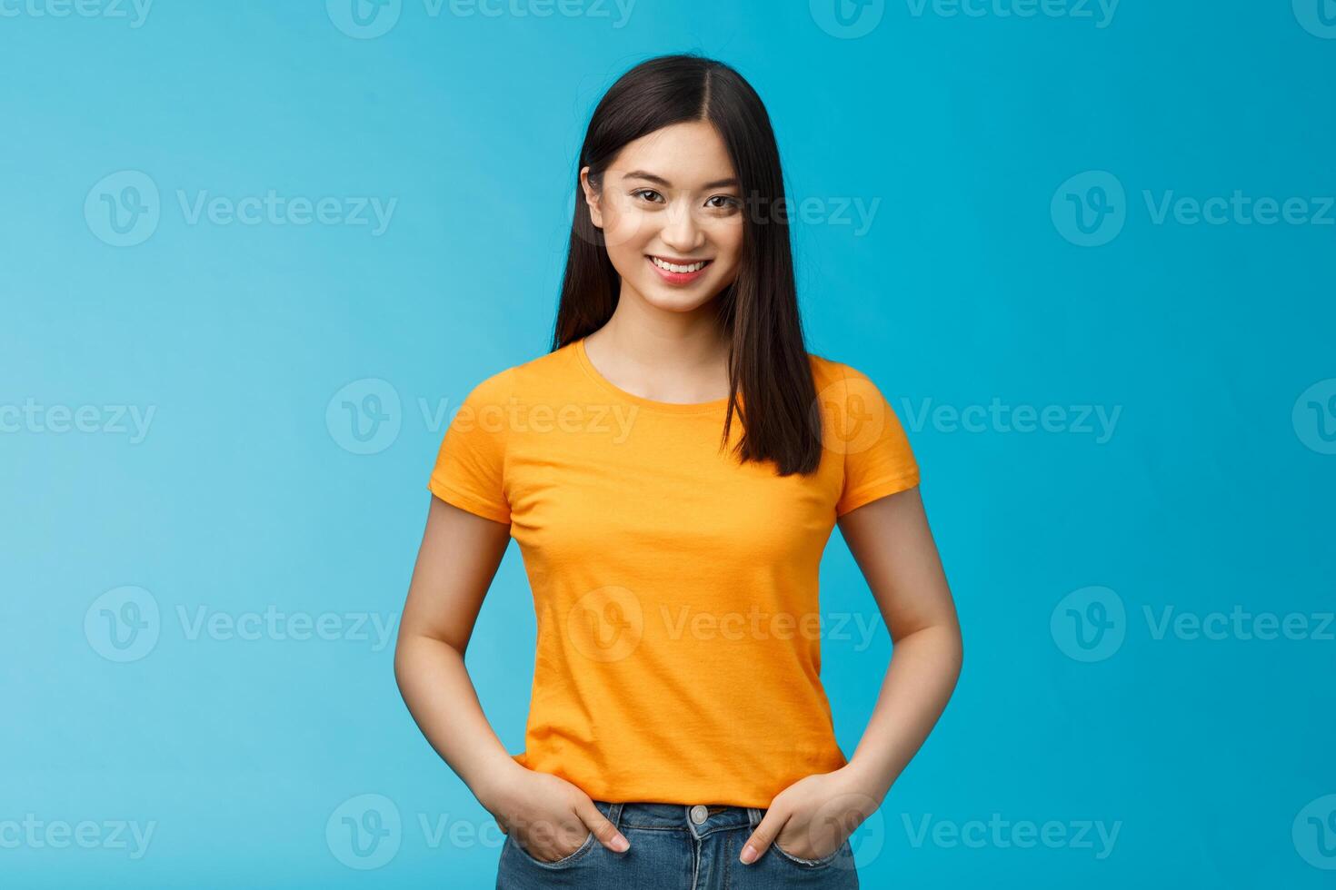 Confident assertive smiling asian woman stand blue background look camera, hold hands jeans pockets express self-assured vibe, enjoy seeing positive results skincare procedures, wear yellow t-shirt photo