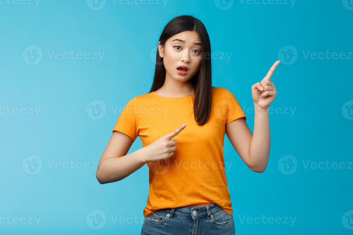 Intrigued curious asian silly tender girl asking question shop assistant opened mouth, questioned interested look, pointing upper right corner puzzled stand blue background photo