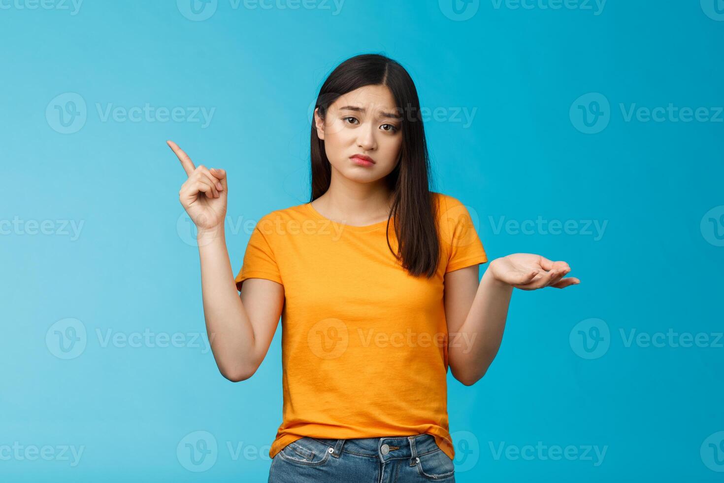 Troubled cute upset asian girl have problem asking your help, pointing upper left corner shrugging unsure, look frustrated and sad, questioned answer, cannot deal problem, stand blue background photo