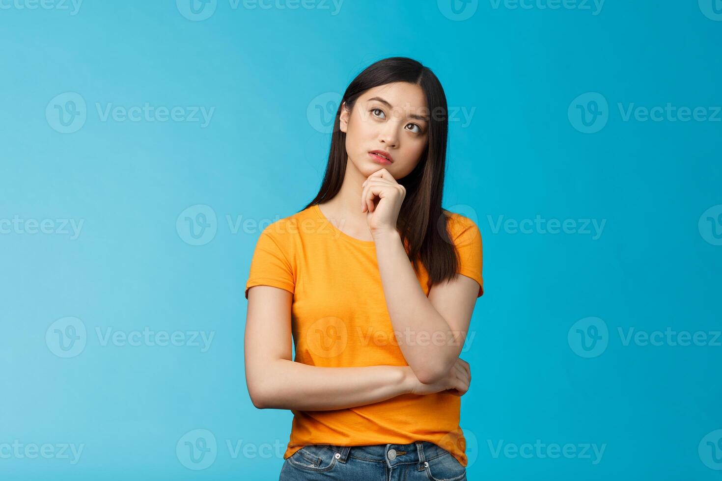 Thoughtful dreamy smart asian girl thinking, making plan inside head, touch chin, look up focused, contemplating interesting scene, pondering, making choice, taking decision, stand blue background photo