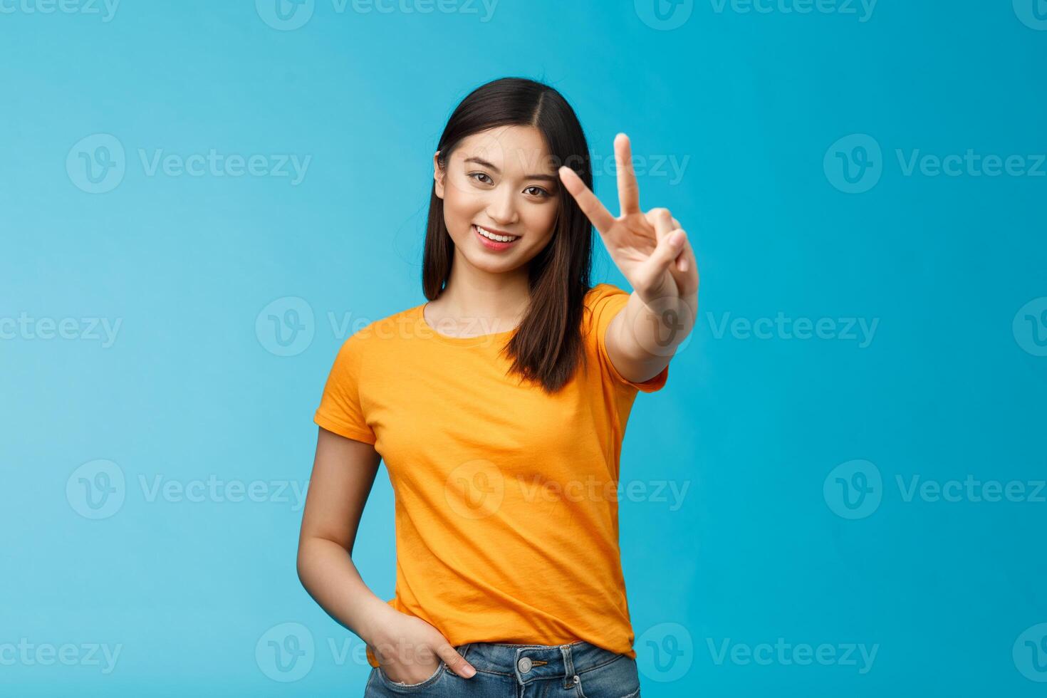 Cheerful friendly asian girl pacifist smiling happily show peace, victory sign, determined win aim achieve success stand blue background joyful, feel luck enthusiastic, wear yellow summer t-shirt photo