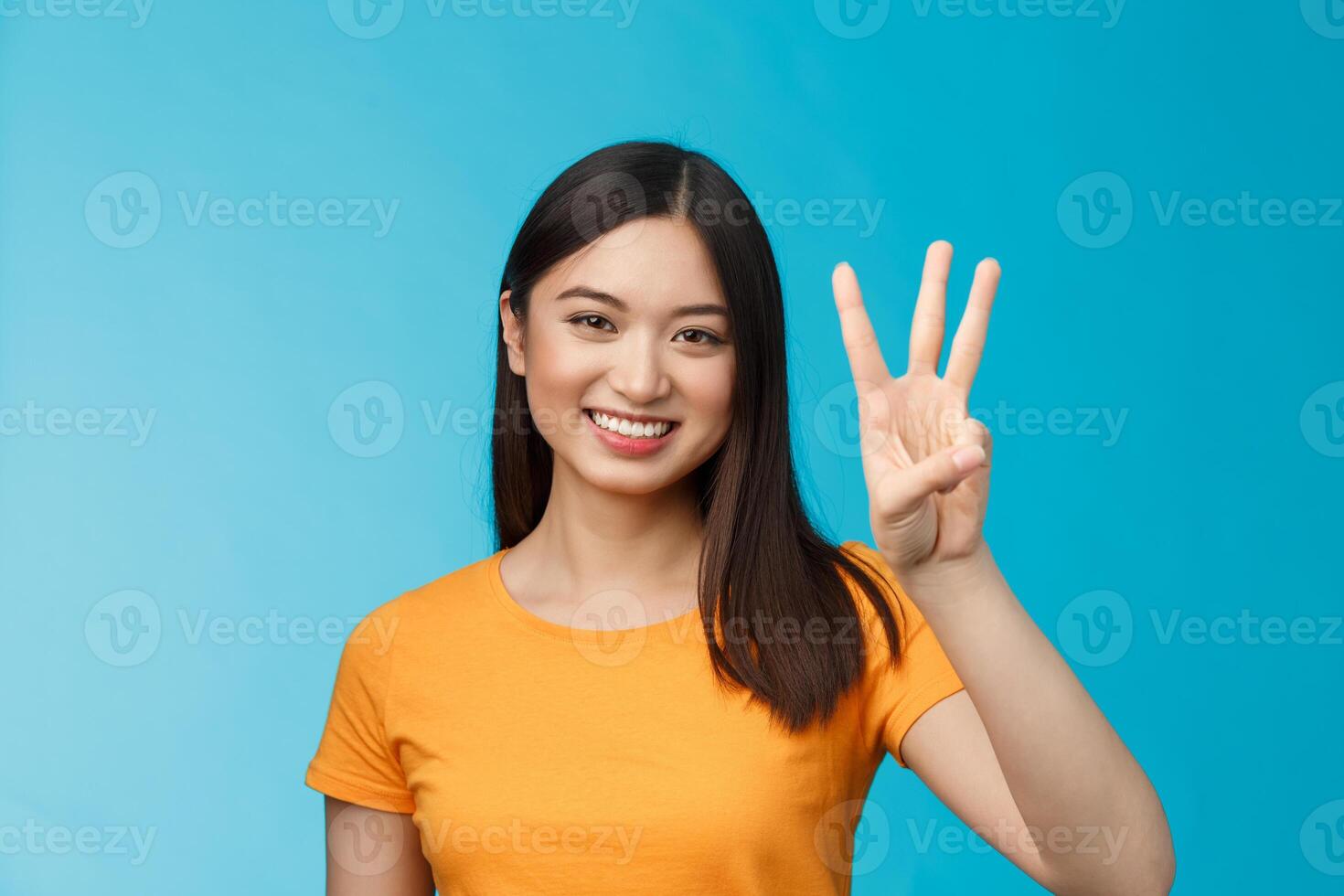 Friendly enthusiastic tender asian woman making reservation for three, number third, smiling broadly, stand blue background joyfully, posing near blue background in yellow t-shirt photo