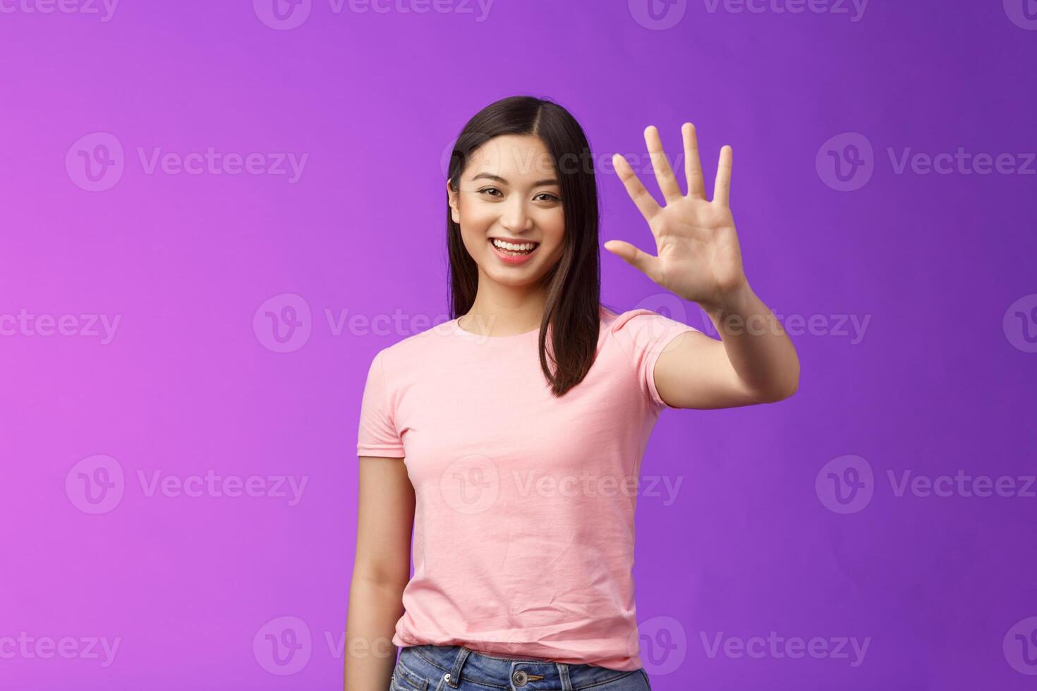 Friendly cute asian tender girl raise palm waving goodbye, smiling joyfully, farewell, gladly welcome friends inviting come in, saying hi hello, greeting, stand purple background photo