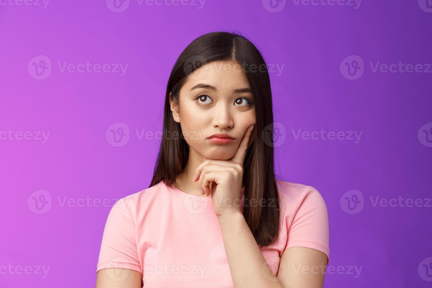 Creative dreamy asian female thinking, have tough decision mind, touch cheek thoughtful, look up, pondering making choice, stand hesitant sighing troublesome thoughts, stand purple background photo