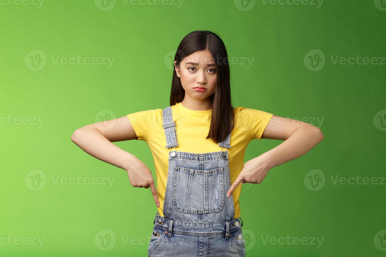 Sad asian girl in dispair feel upset, frowning sulking pessimistic, pointing down look camera pulling sorrow uneasy face, express sadness and frustration, stand green background photo