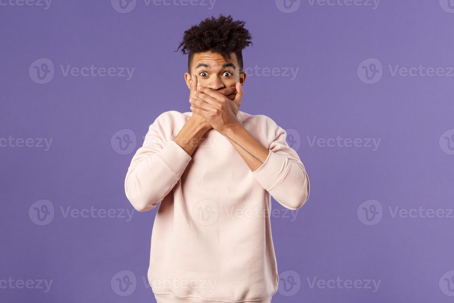 Portrait of shocked impressed young hispanic man found out gossip, heard rumour, trying not laugh shut his mouth with hands and express amazement and surprised with gaze, purple background photo