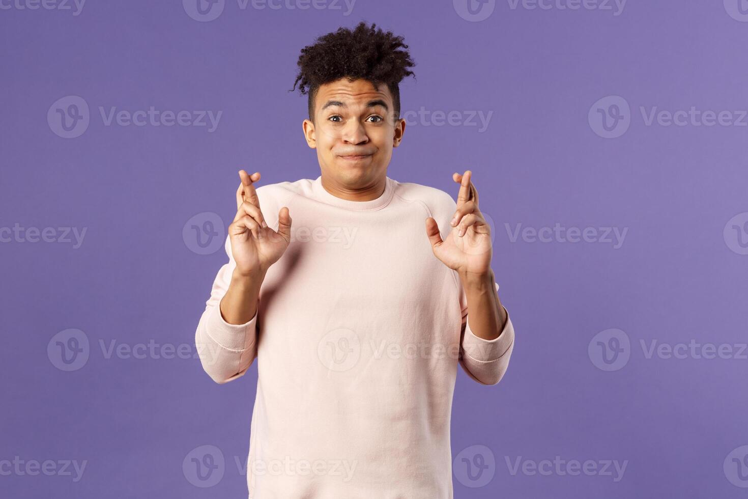 Portrait of young hopeful handsome man with dreads ancitipating something good happen, cross fingers good luck, smiling with joy and optimism, having faith, praying over purple background photo