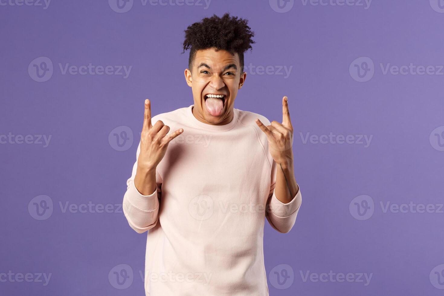 Portrait of enthusiastic, excited funny young man having fun, enjoying awesome rock concert, show heavy metal sign, stick tongue and rejoicing hear favorite band on stage, purple background photo
