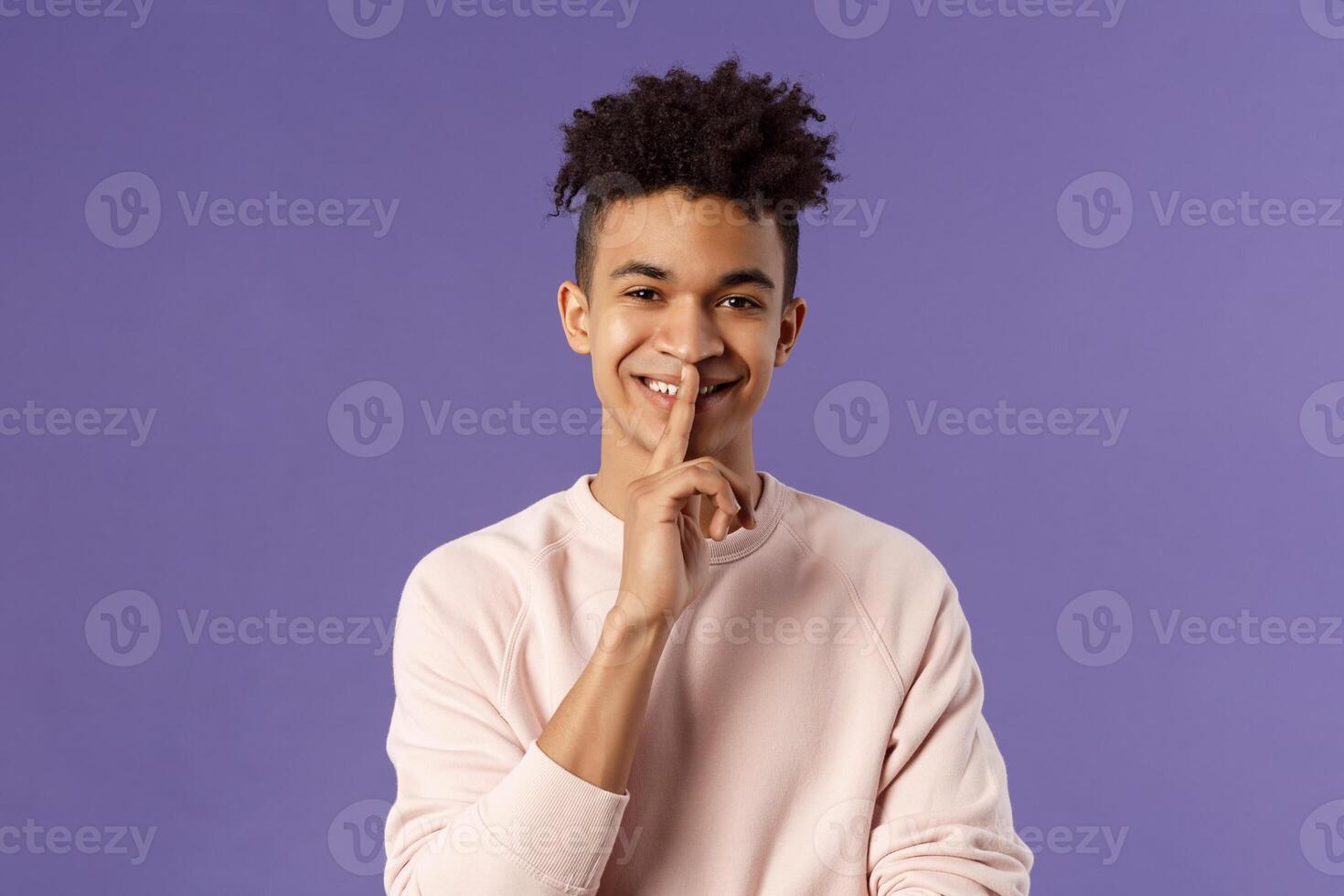 Close-up portrait of attractive smiling young man hiding secret, asking keep silent or quiet to do surprise, show shush gesture, place index finger to mouth, standing purple background photo