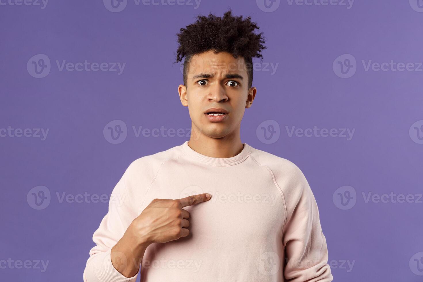 Close-up portrait of embarrassed and worried young man being named or called to principal office, pointing at himself and asking why with disbelief and anxiety, standing purple background photo