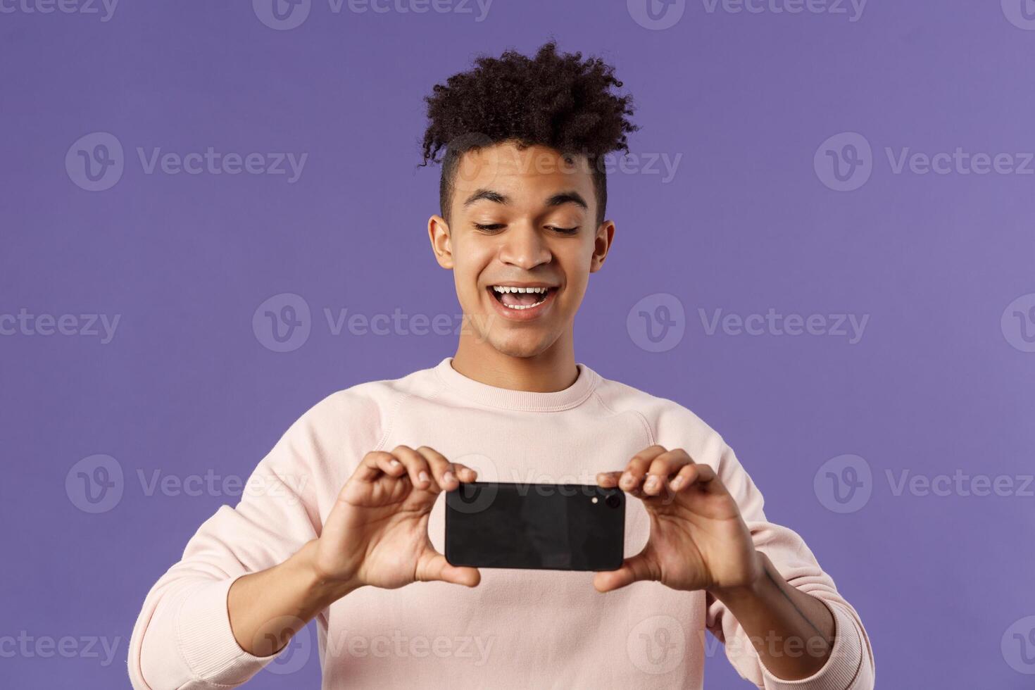Portrait of amazed, excited young man seeing something interesting, stream concert to his internet social network profile, taking photo or recording with mobile phone, purple background