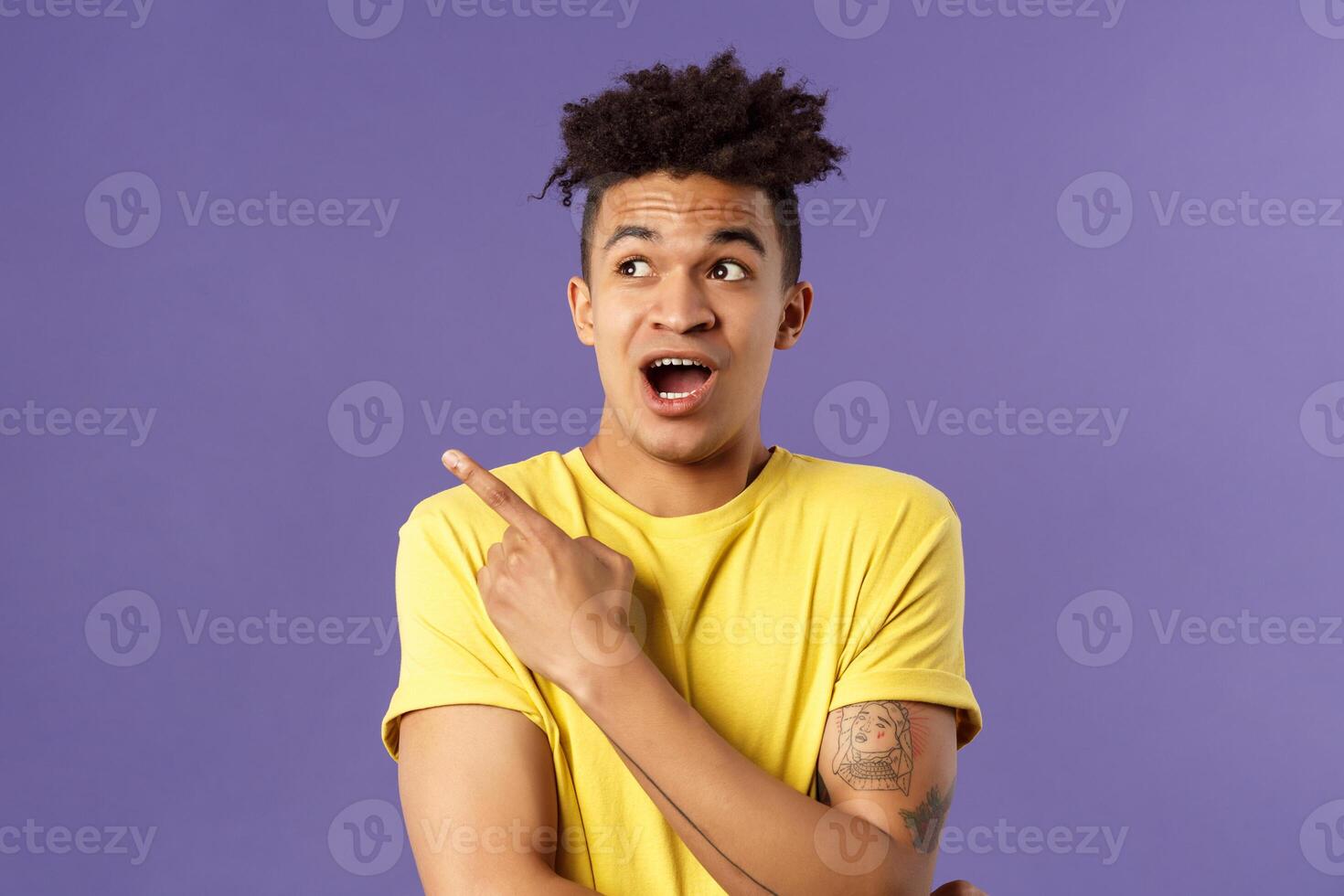 Portrait of impressed hispanic guy seeing something interesting, pointing finger and looking upper left corner, discuss strange thing upwards, standing purple background curious photo