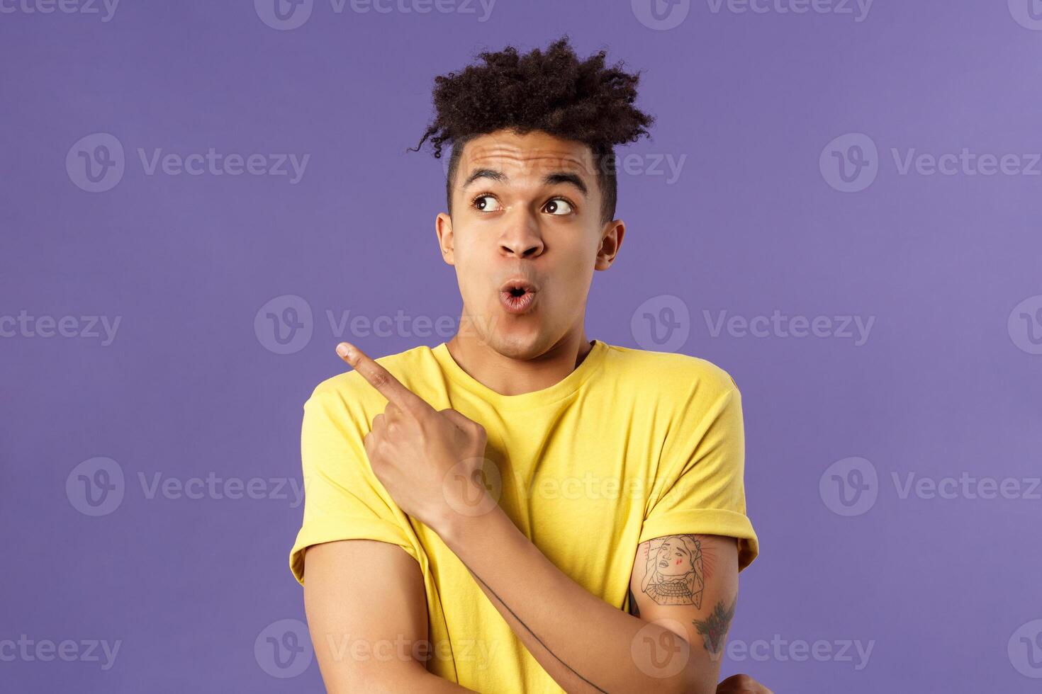 Waist-up portrait of amazed, impressed hispanic male with dreads, seeing something incredible and cool, pointing finger looking upper left corner, breathtaking awesome thing, purple background photo