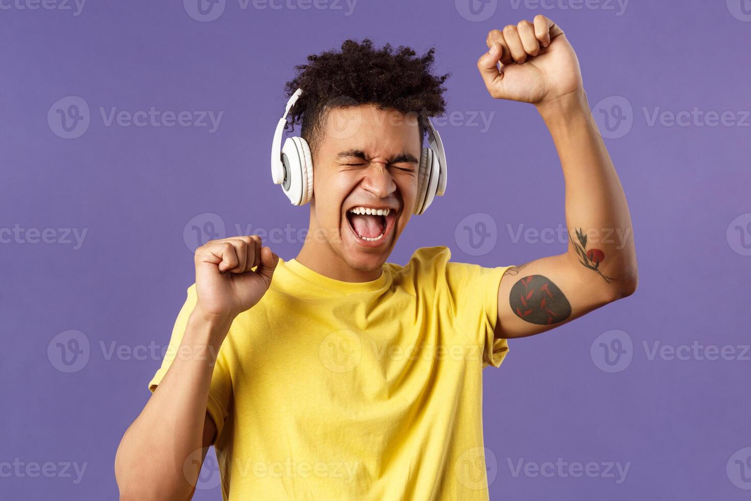 Close-up portrait of cheerful, happy young dancing guy lift hand up singing along, close eyes and smiling upbeat as listening awesome song in headphones, enjoying music, purple background photo