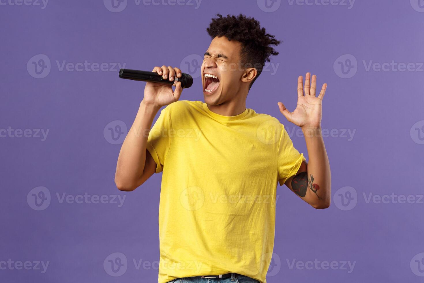 Portrait of passionate carefree young hispanic singer with dreads and tattoos, reaching highest note in song, raising hand up singing loud at microphone with closed eyes, purple background photo
