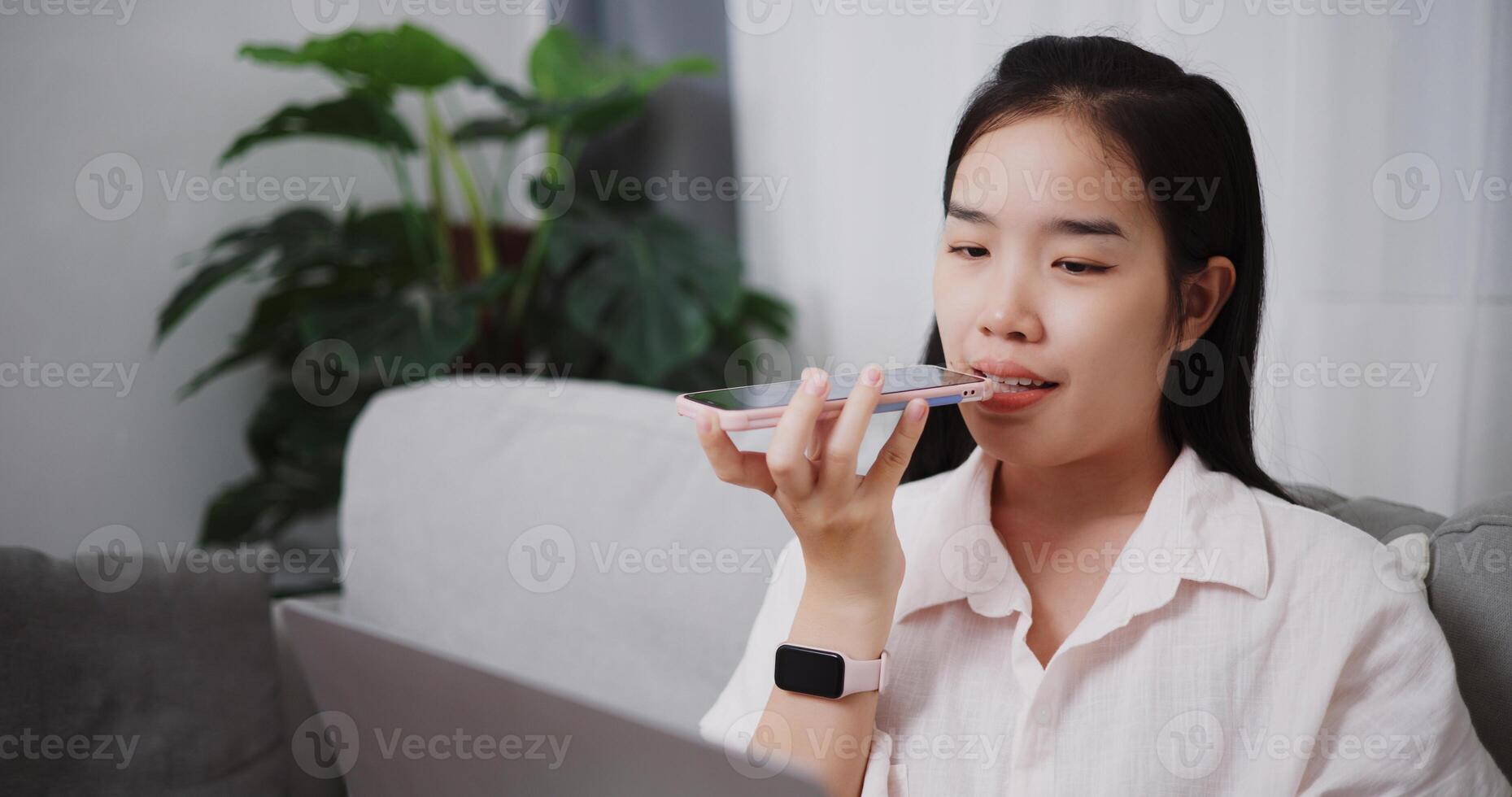 Portrait of Young asian woman recording a message on cell phone while working on laptop in the living room at home ,Lifestyle with device technology, Working from home photo