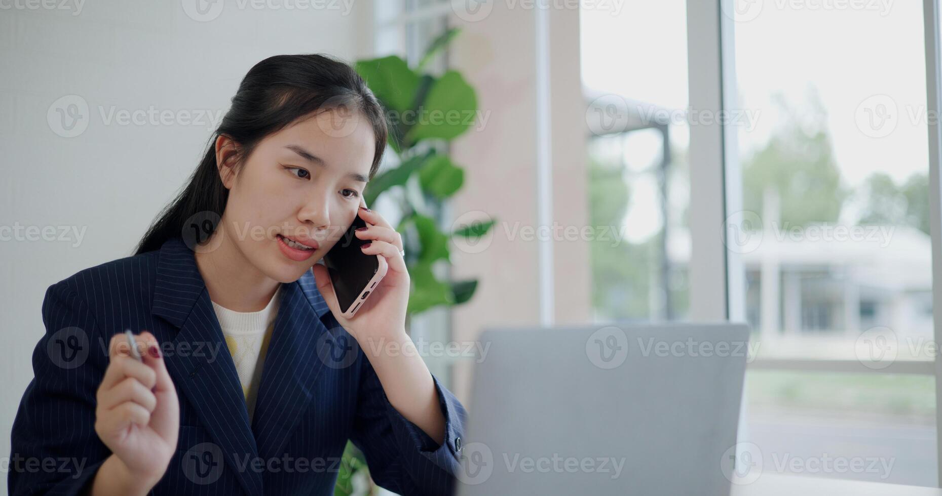 Portrait of Asian businesswoman in suit using mobile phone to explain work to colleagues while working on laptop at small coffee shop. Work from anywhere, Remote working photo